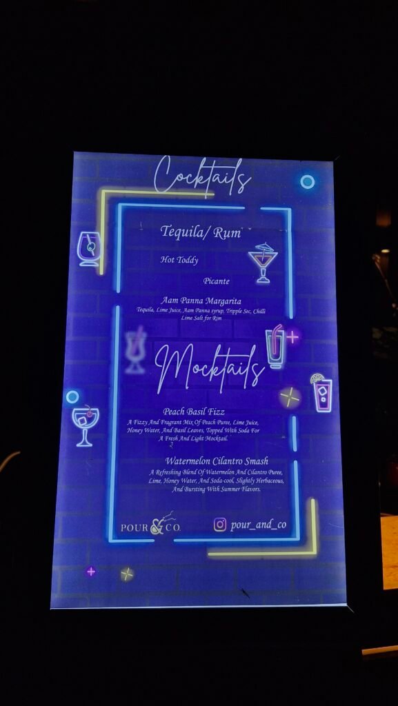 pre-wedding bar setup for an outdoor Haldi pool party. A guest muddles fresh ingredients into a signature coupe glass at the interactive 'Muddle-Your-Own-Botanical' station.